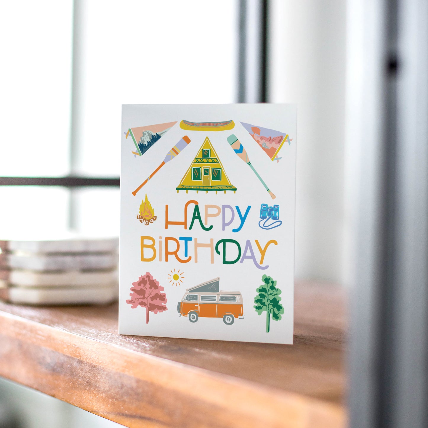 A colorful camping-themed birthday card featuring an A-frame cabin and a camper van. Perfect for outdoor lovers and adventurers.