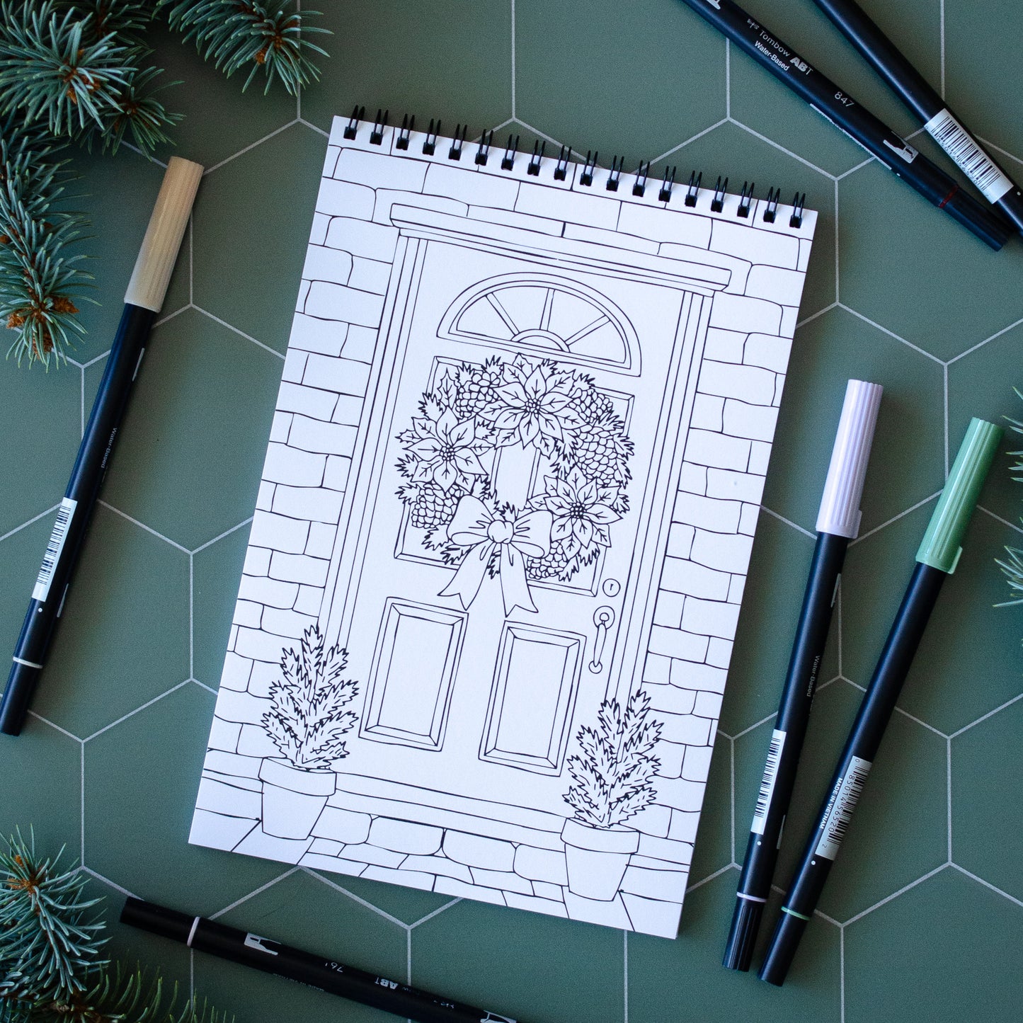 Coloring book page with a door design on a green surface with markers and pine branches.