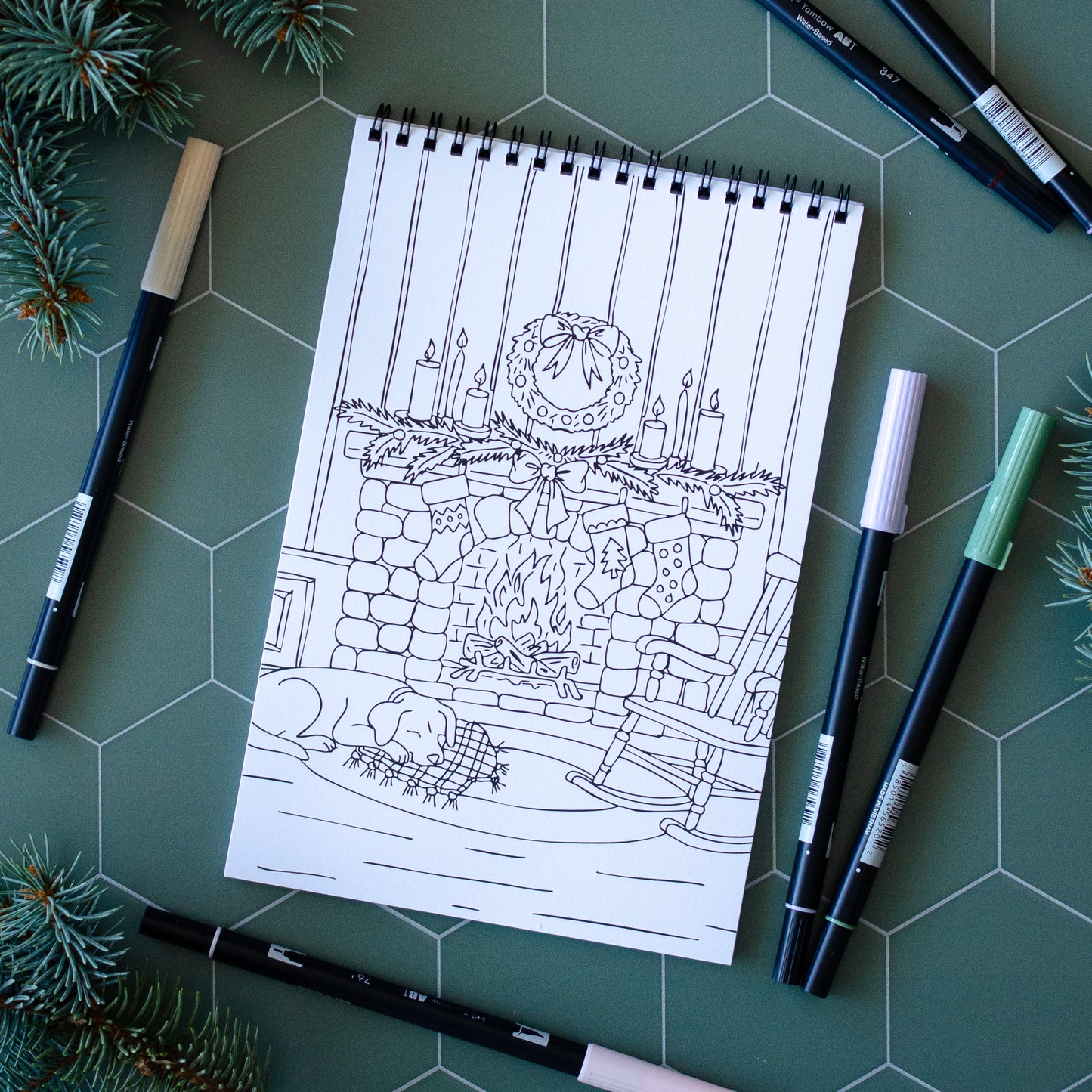 Coloring book page with a Christmas-themed illustration on a green surface with markers and pine branches.