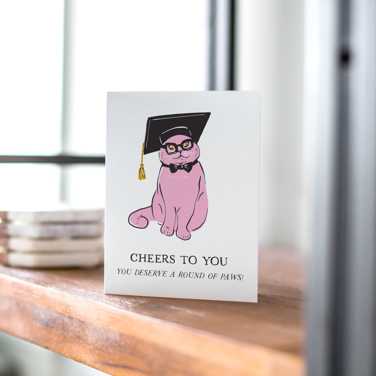 A stylish greeting card featuring a pink cat wearing a black graduation cap, glasses, and a bow tie. The card includes the message "Cheers to You" with the pun, "You deserve a round of paws!" Perfect for celebrating graduations and academic achievements.