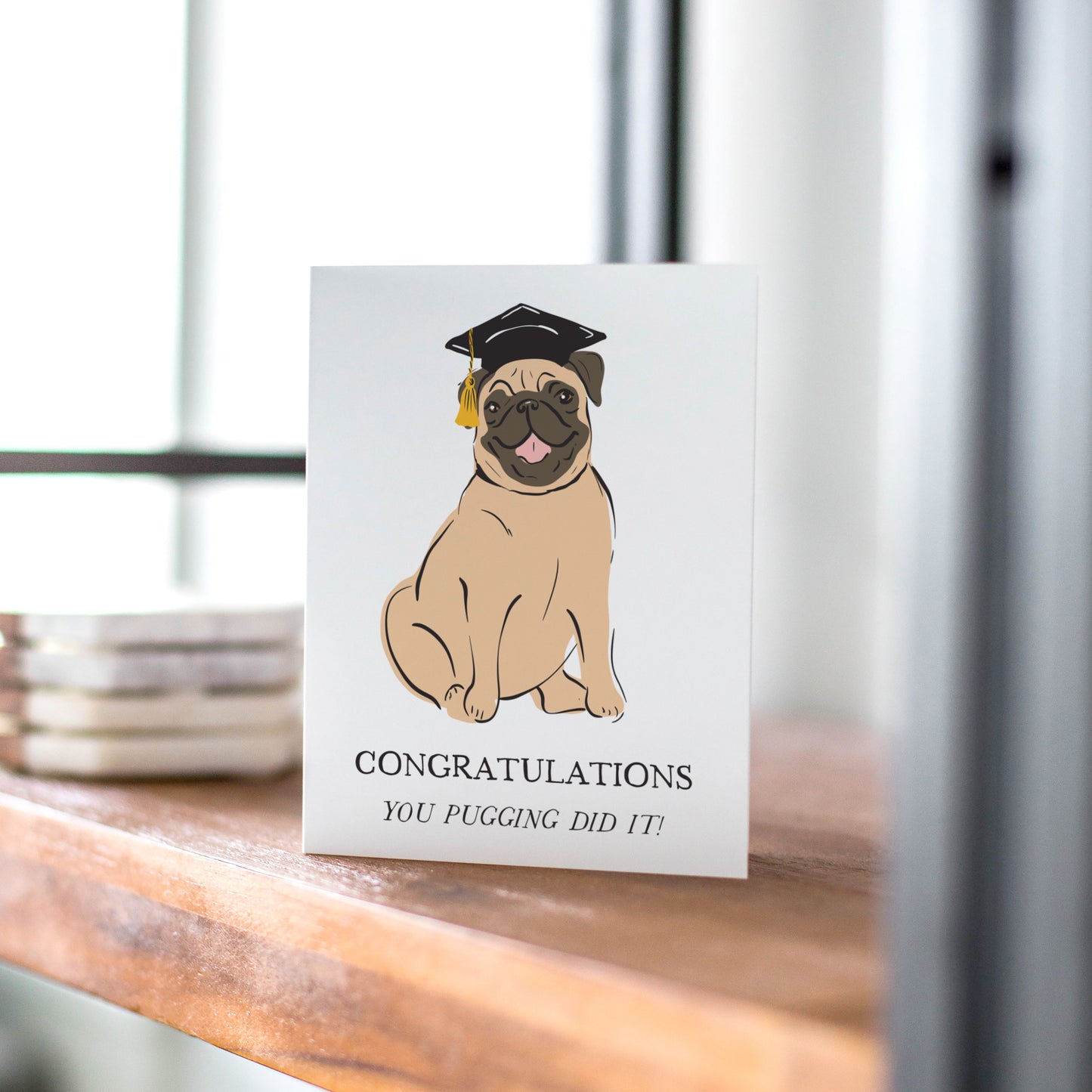 A fun and adorable greeting card featuring a happy pug wearing a black graduation cap with a gold tassel. The card displays the message "Congratulations" with the playful pun, "You pugging did it!" Perfect for celebrating graduations and academic milestones