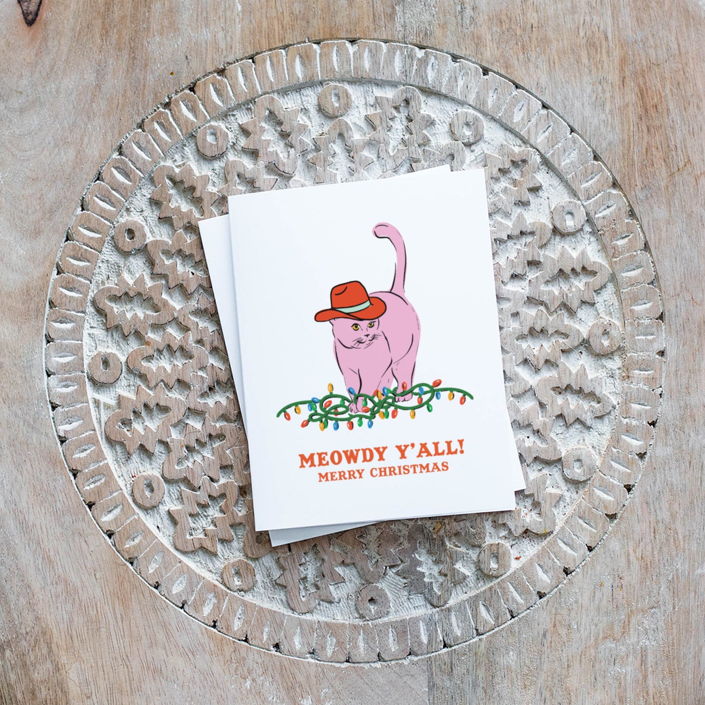 Christmas card with a pink cat wearing a red cowboy hat sitting on Christmas lights and the text “Meowdy Y’all! Merry Christmas.”