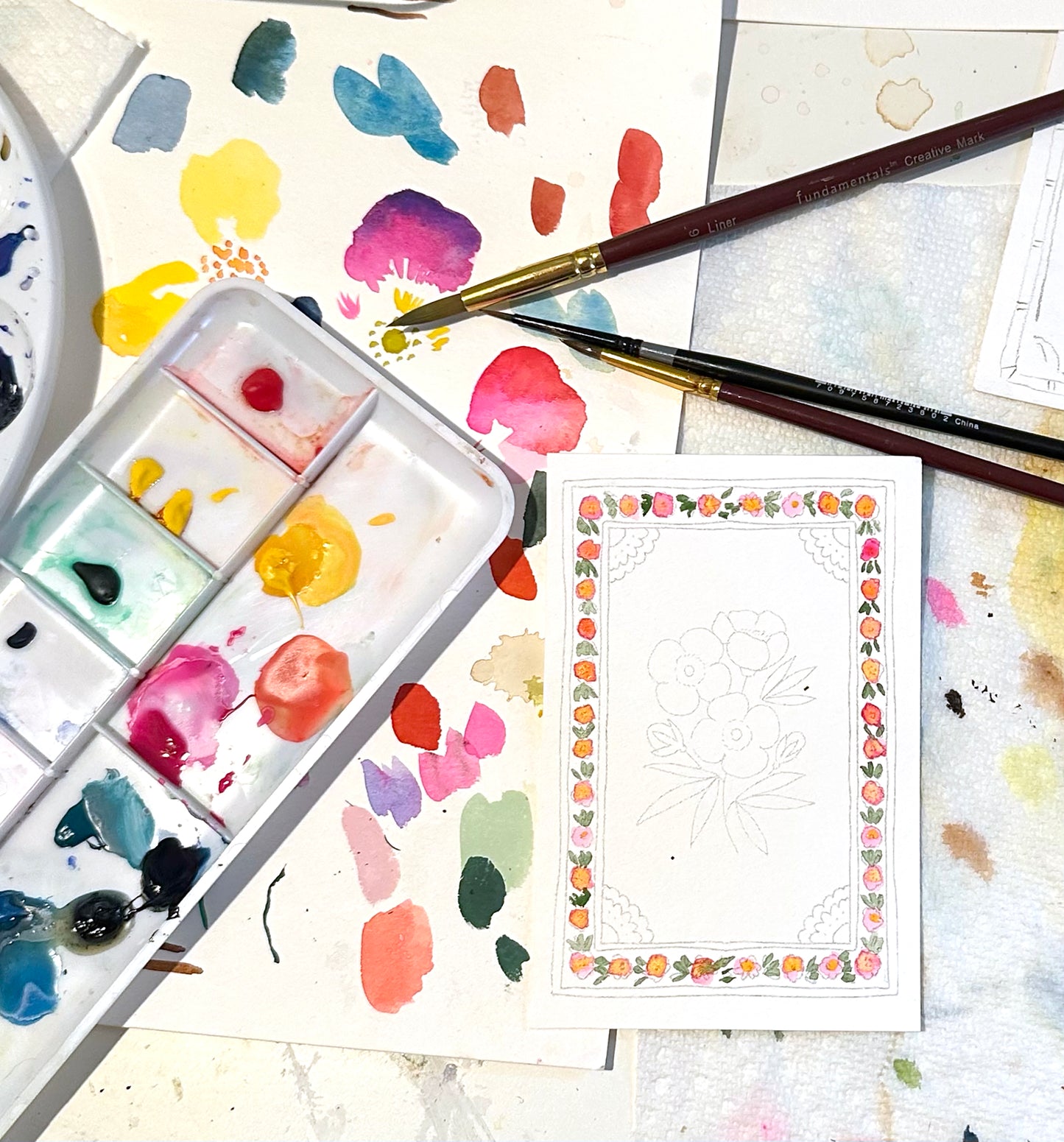 Artistic setup with watercolor paints, brushes, and a partially completed watercolor card on a textured surface.