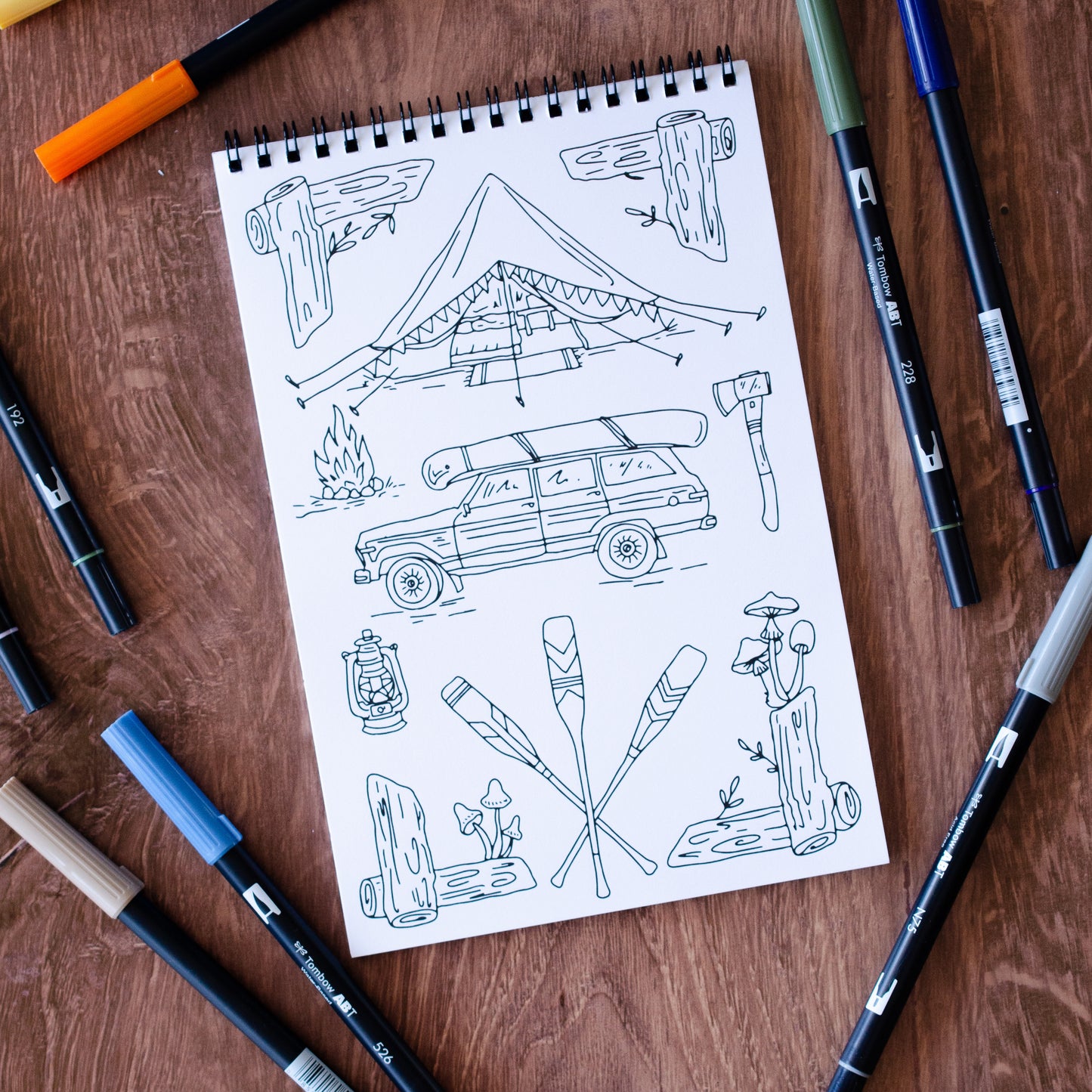 The Great Outdoors Coloring Book with 22 pages of hand-drawn nature illustrations featuring a camping scene, tent, pine trees, wildlife, and a cozy campfire under the stars. Spiral-bound adult coloring book for outdoor and adventure lovers.