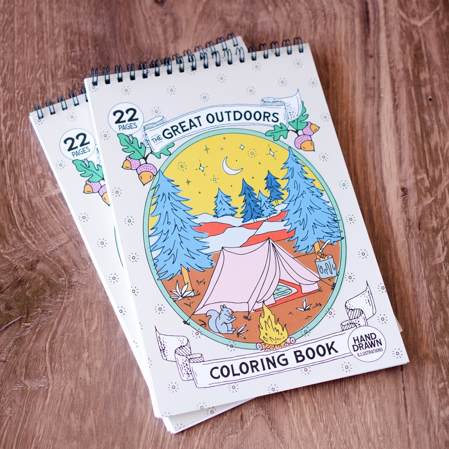 The Great Outdoors Coloring Book with 22 pages of hand-drawn nature illustrations featuring a camping scene, tent, pine trees, wildlife, and a cozy campfire under the stars. Spiral-bound adult coloring book for outdoor and adventure lovers.