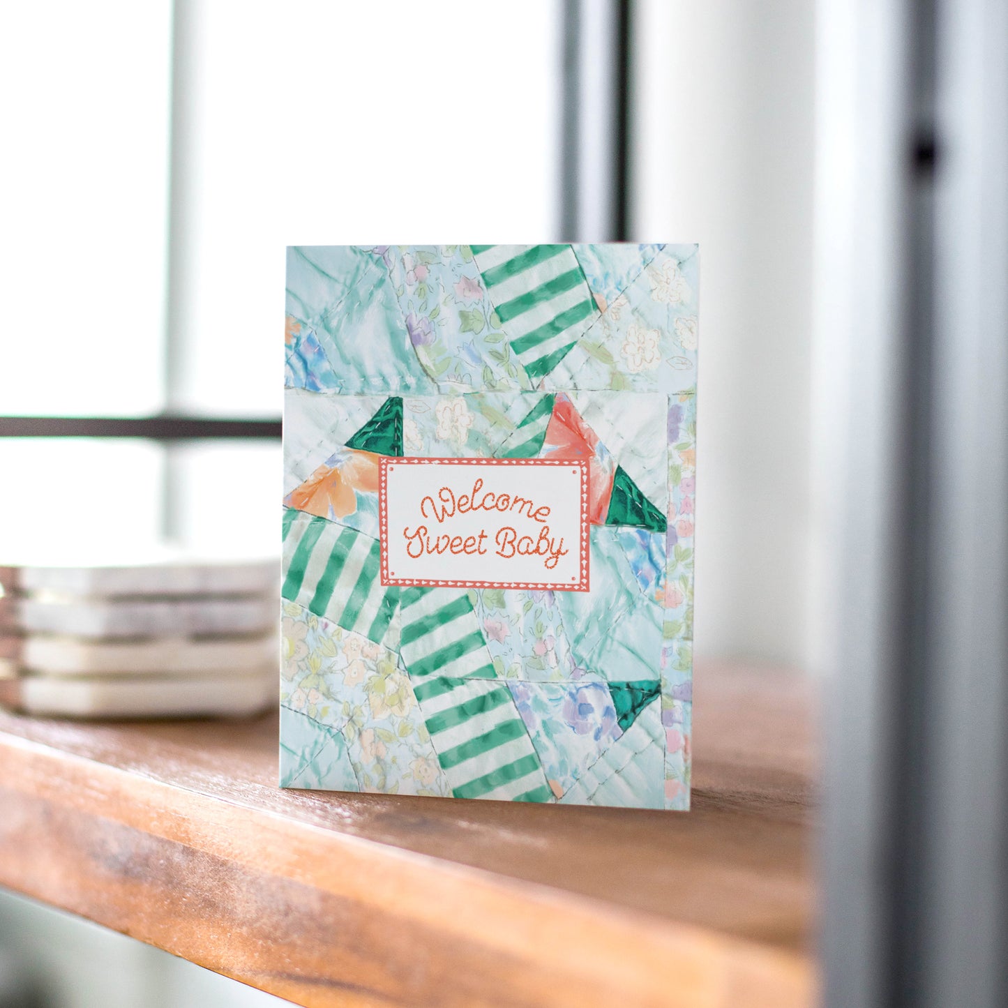 A charming greeting card featuring a quilt-inspired background with soft, patchwork patterns. The design includes a quilt tag with the message "Welcome Sweet Baby," making it a heartfelt choice for celebrating a new arrival. Perfect for baby showers and newborn gifts.