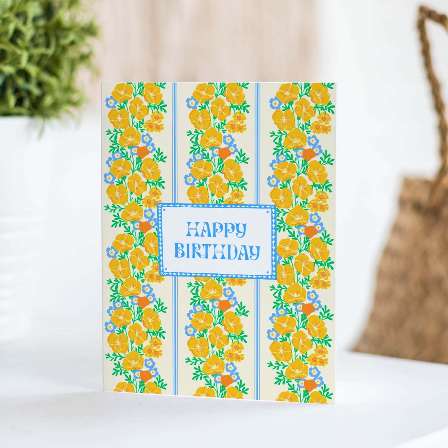 Birthday card with vertical stripes of yellow poppies and blue flowers surrounding “Happy Birthday” text in a blue frame.
