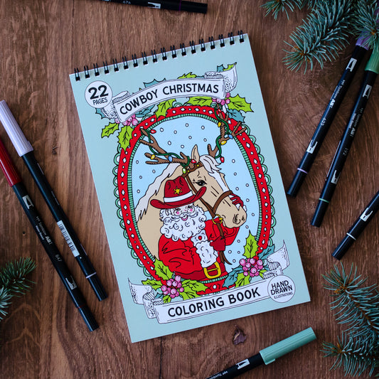 Coloring book titled 'Cowboy Christmas' with markers on a wooden surface