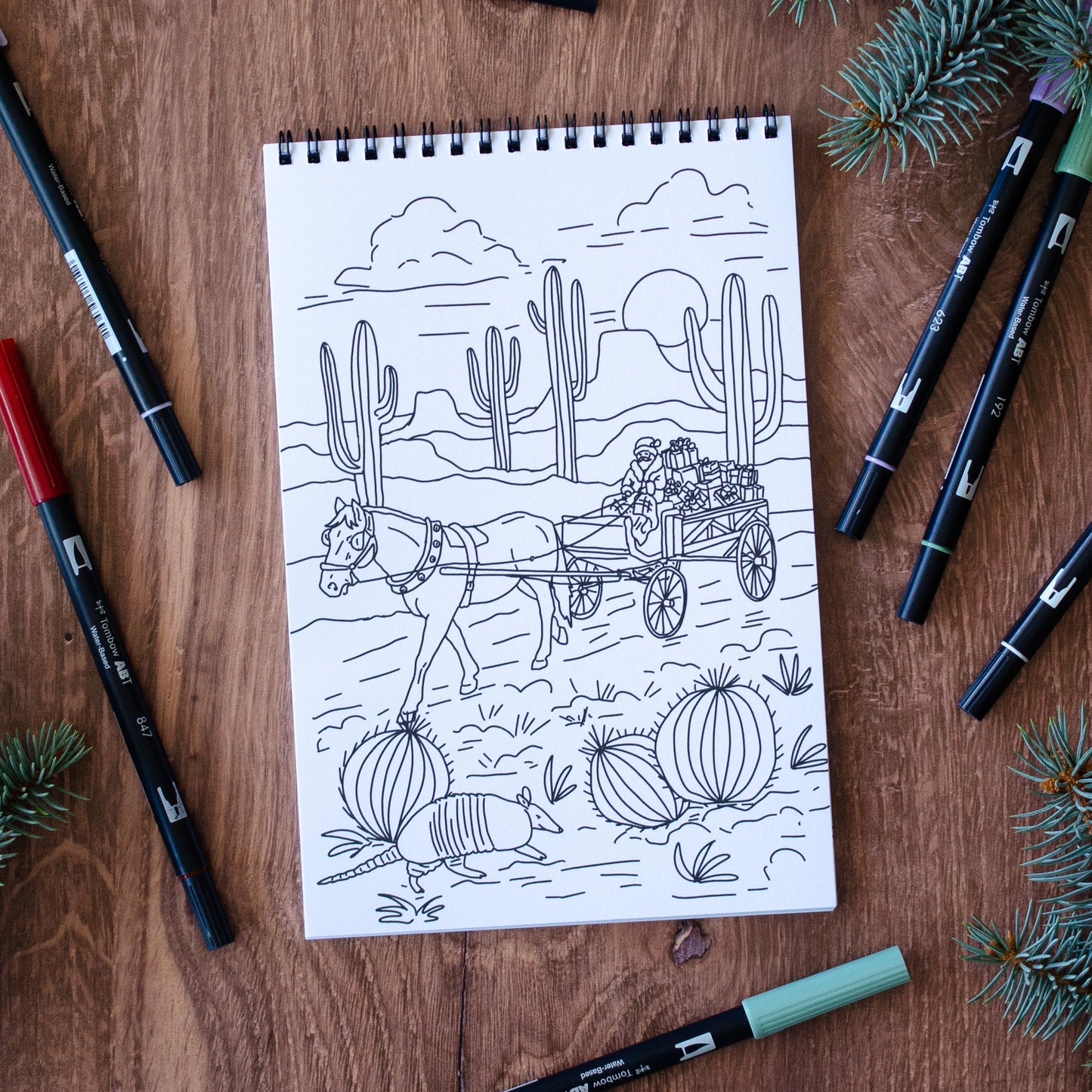 Coloring book page with a desert scene featuring a horse and cart, surrounded by markers on a wooden surface.