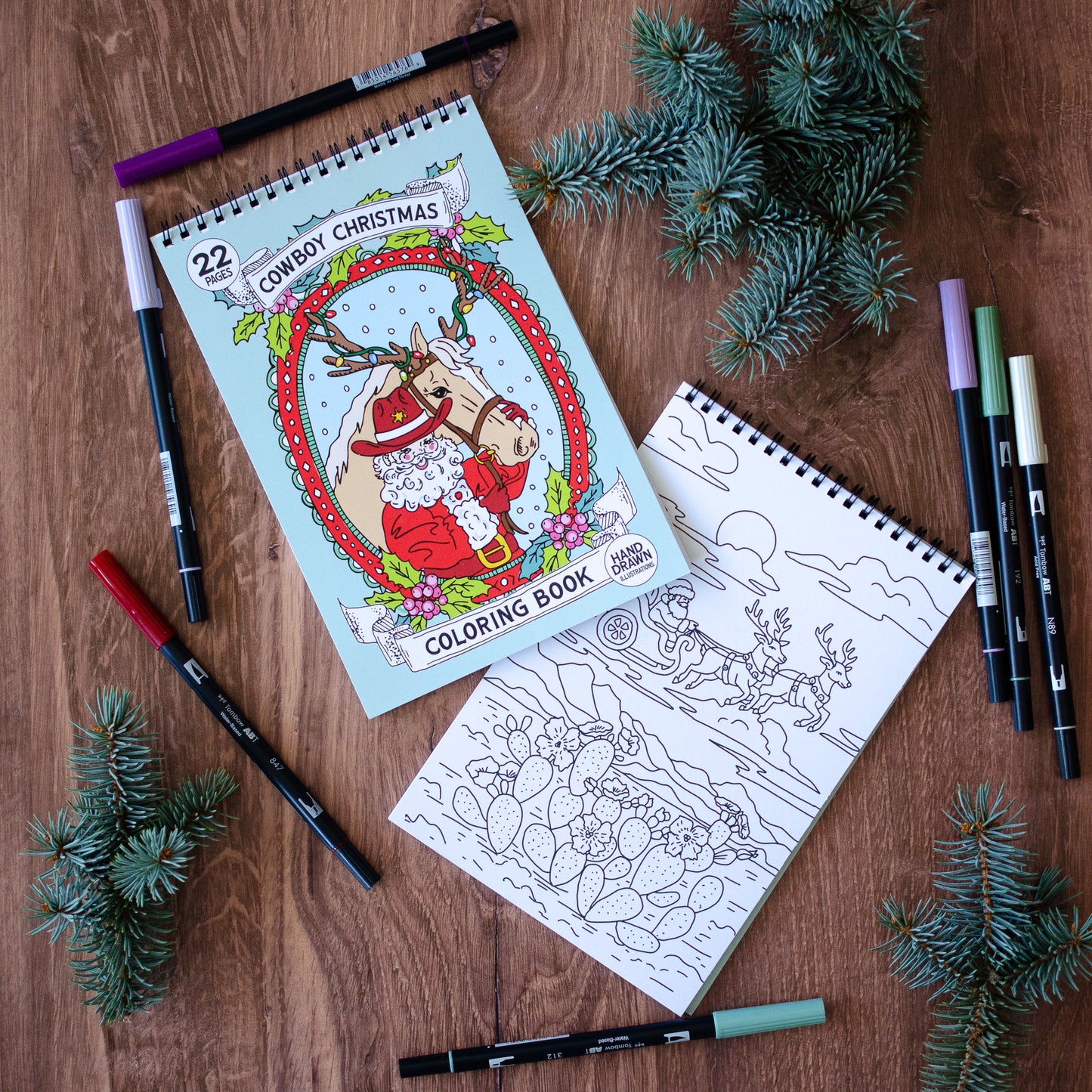 Coloring book with Christmas theme on a wooden surface with markers and pine branches.