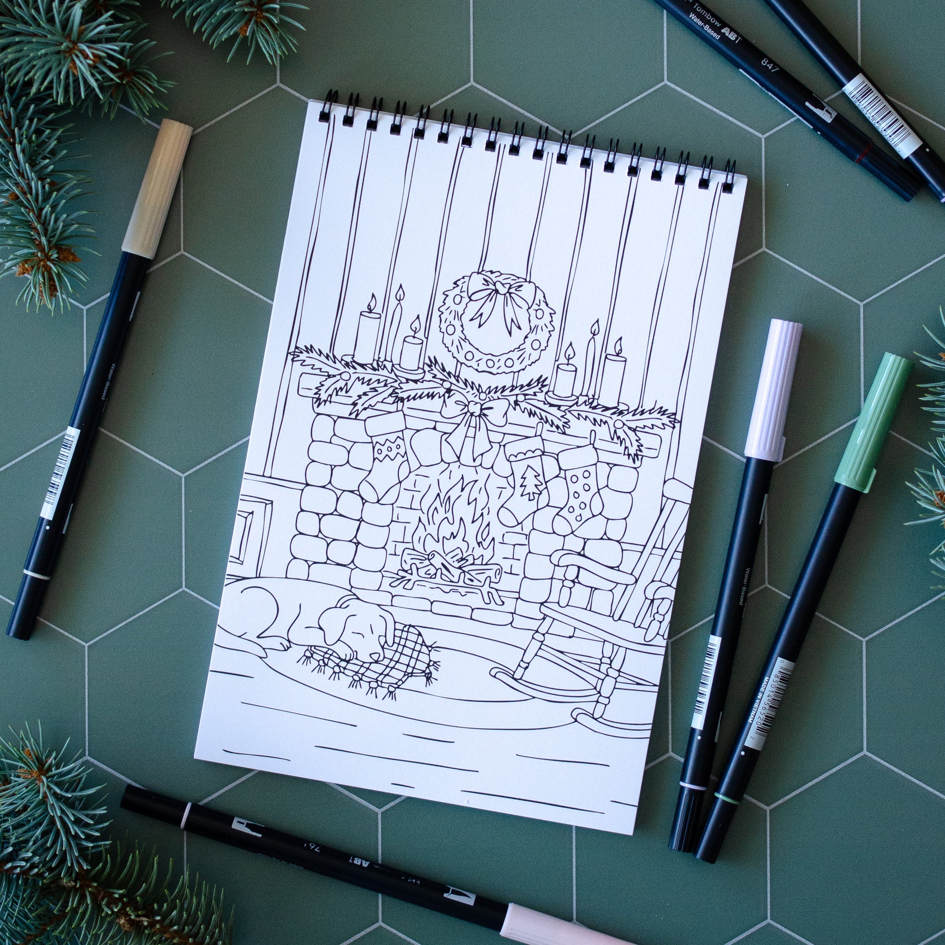 Coloring book page with a Christmas-themed illustration on a green surface with markers and pine branches.