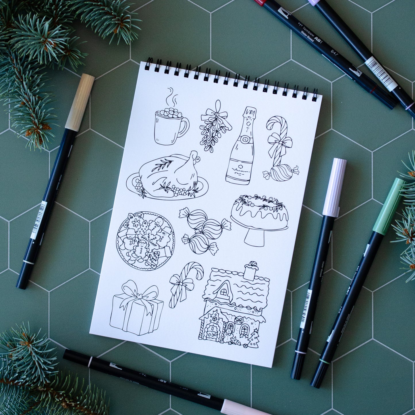 Coloring book page with black line art on a green surface with markers and pine branches.