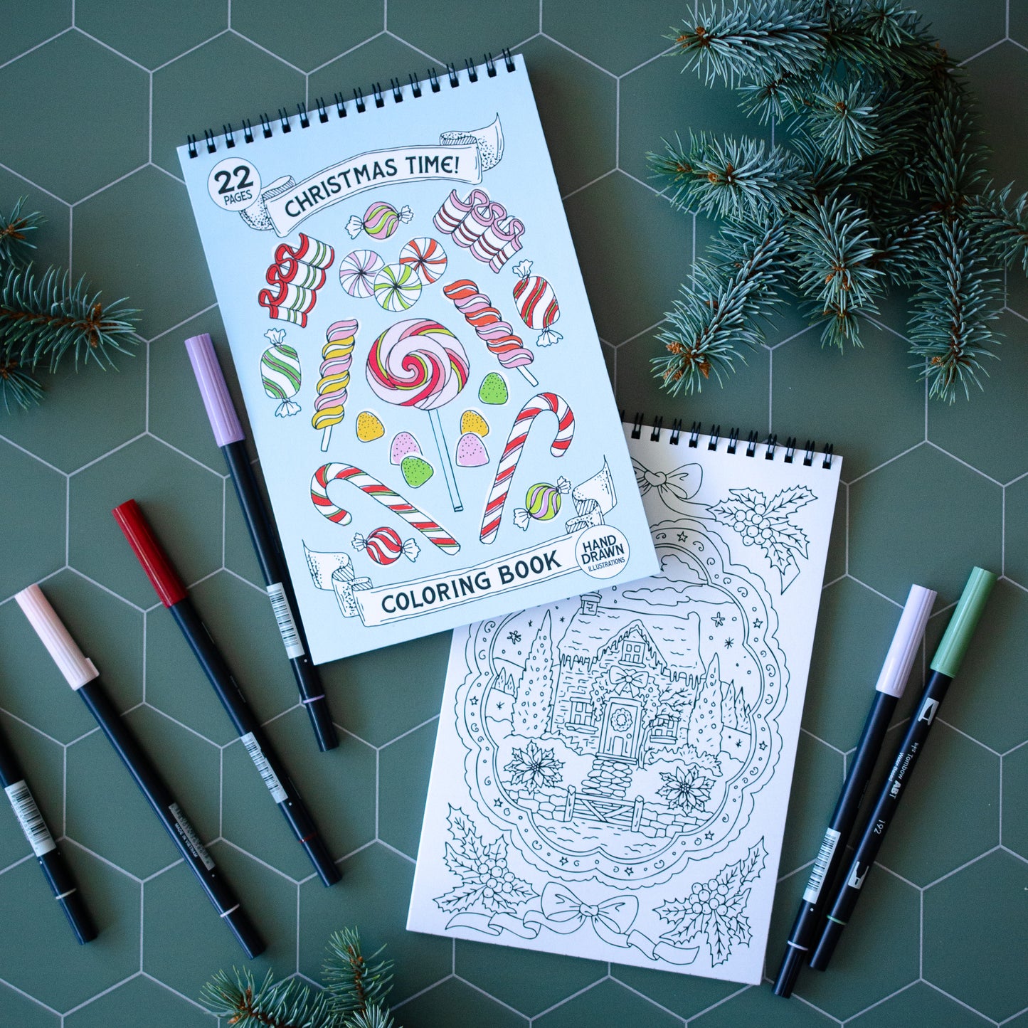 Coloring books with Christmas-themed pages and coloring pencils on a green surface with pine branches.