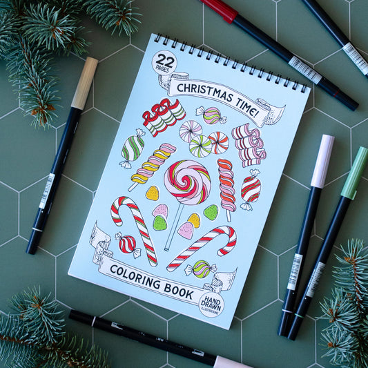 Coloring book titled 'Christmas Time!' with candy illustrations on a green surface with markers and pine branches.