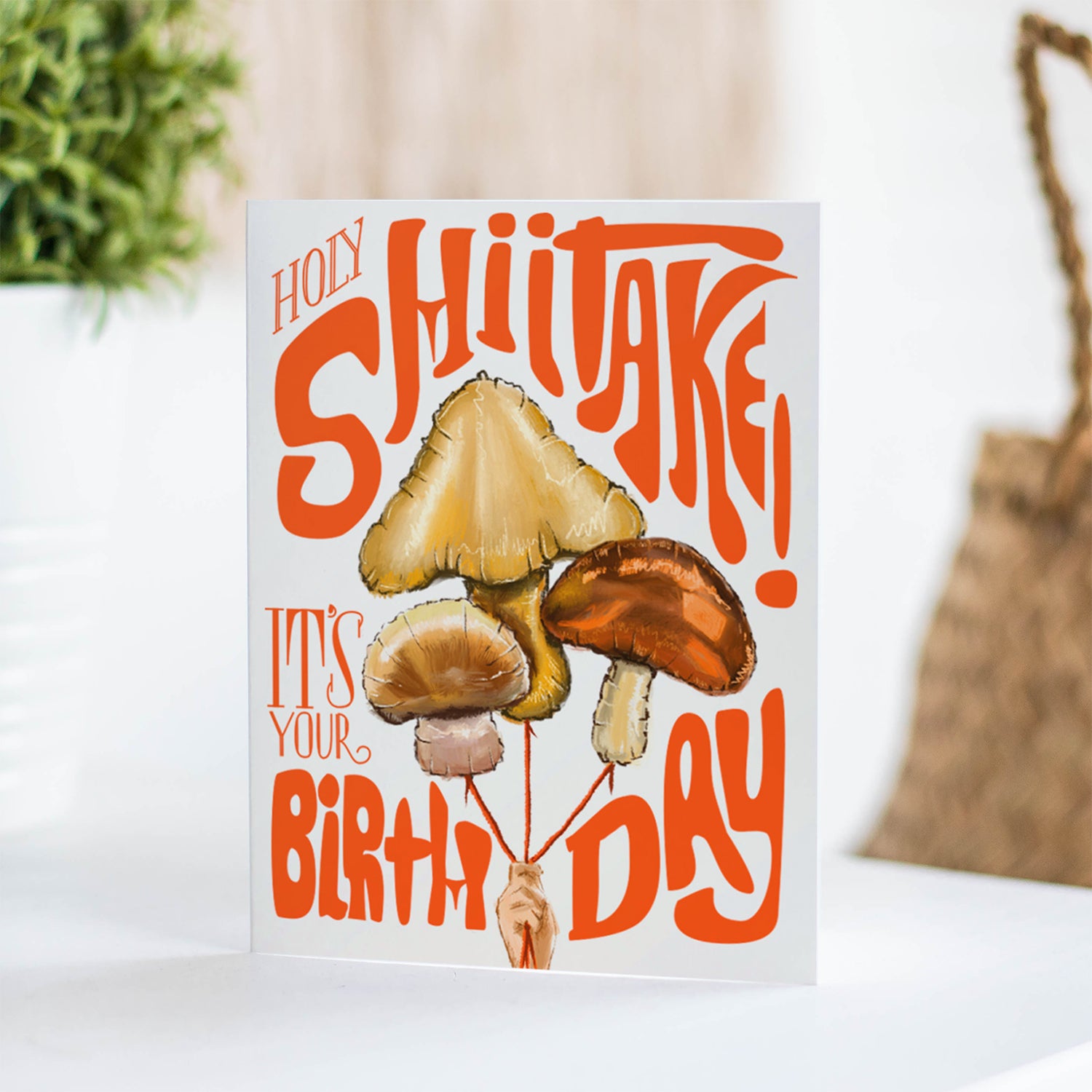 Humorous birthday card featuring a playful mushroom balloons with the phrase 'Holy Shiitake,' perfect for a lighthearted celebration.