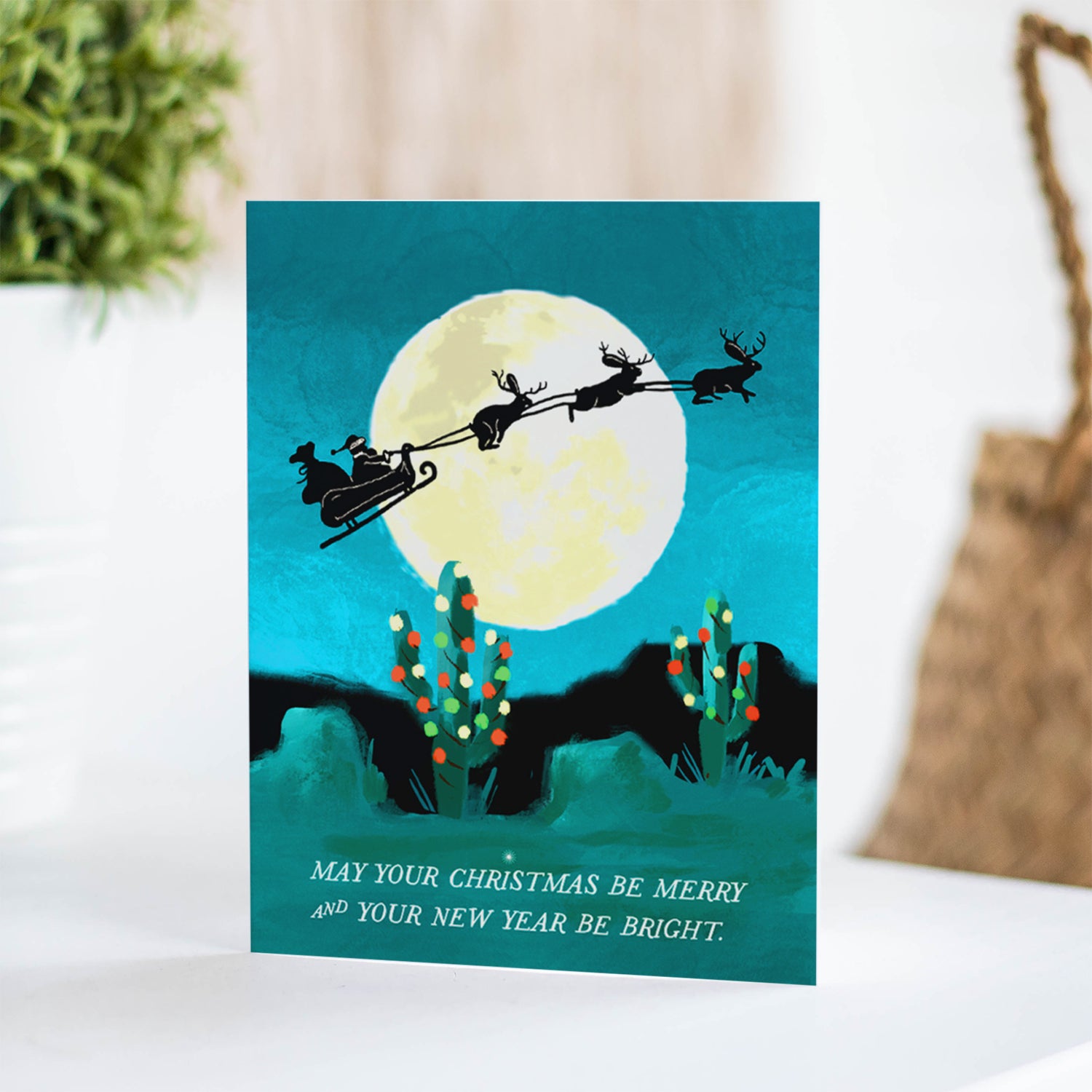 Jackelope Santa holiday card featuring jackelopes pulling Santa's slay across a full moon over a desert landscape, ideal for a unique holiday greeting, blank inside.