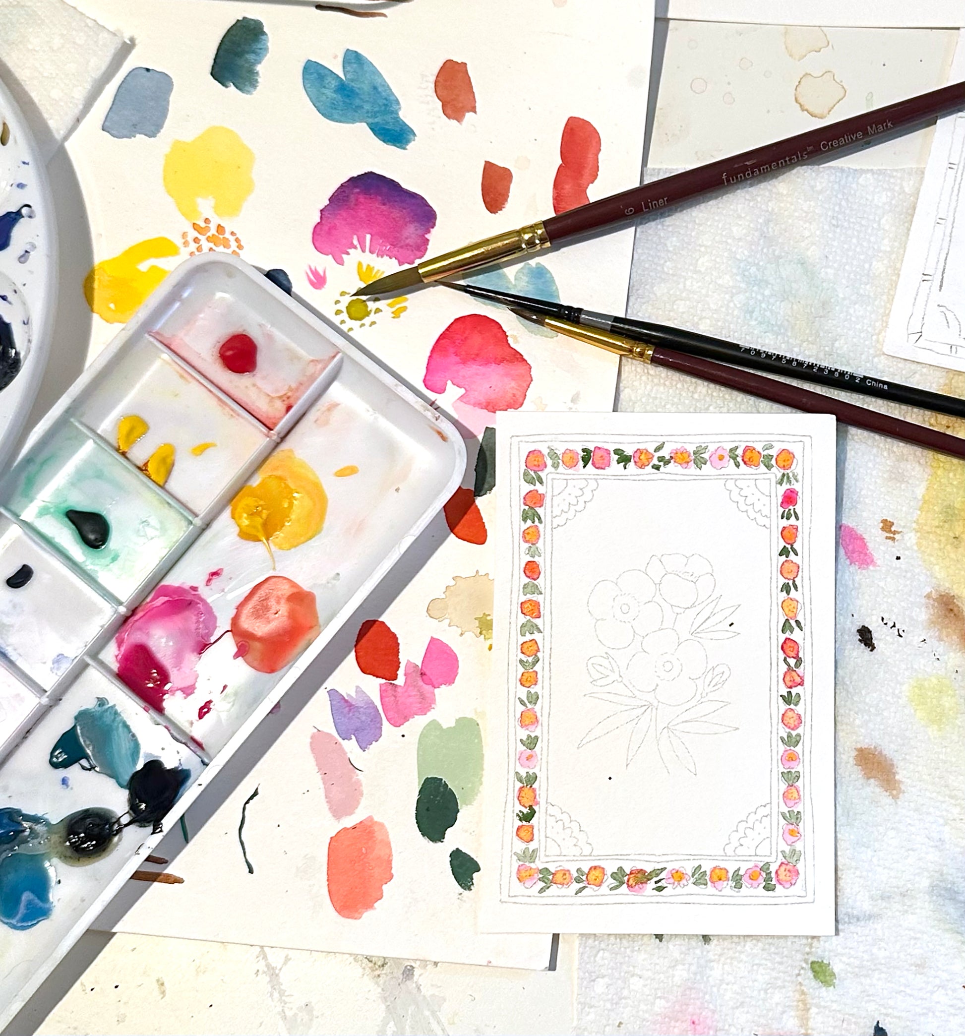 Artistic setup with watercolor paints, brushes, and a partially completed watercolor card on a textured surface.