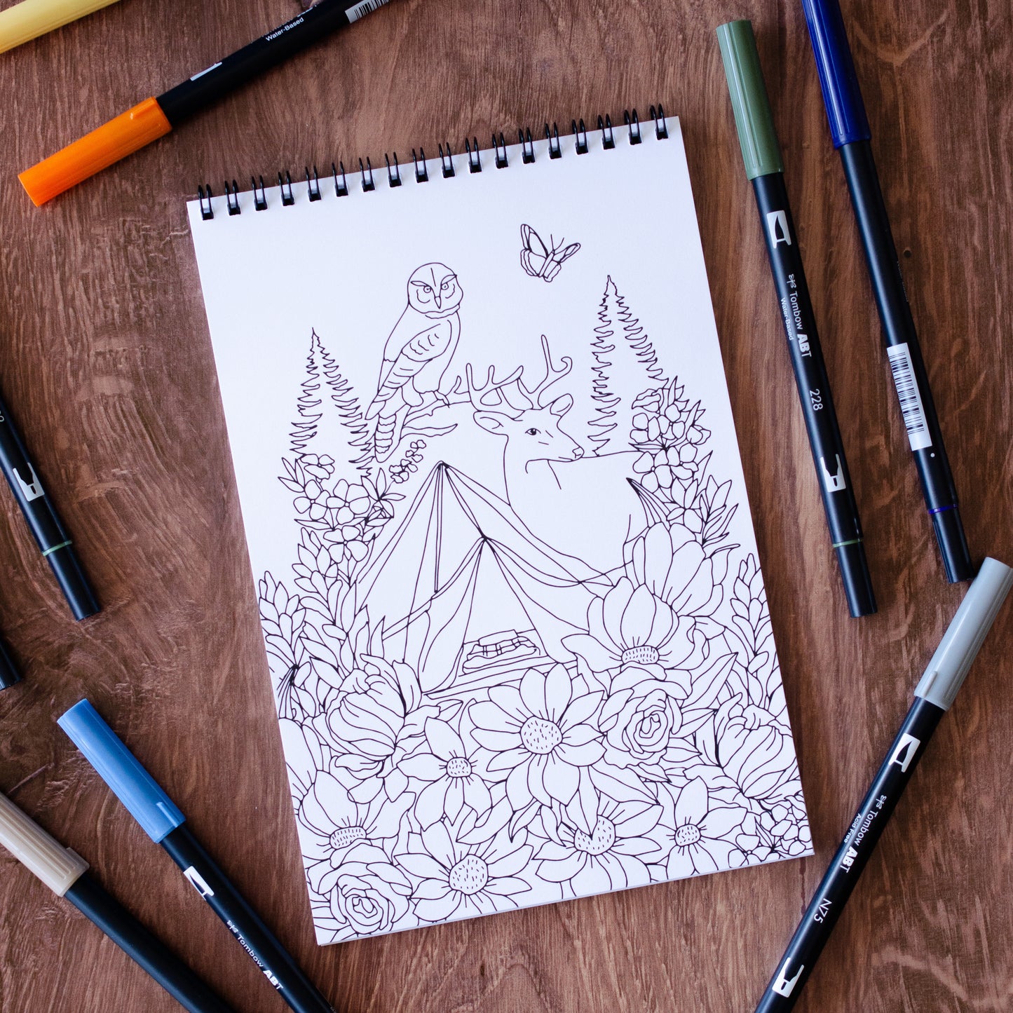 The Great Outdoors Coloring Book with 22 pages of hand-drawn nature illustrations featuring a camping scene, tent, pine trees, wildlife, and a cozy campfire under the stars. Spiral-bound adult coloring book for outdoor and adventure lovers.