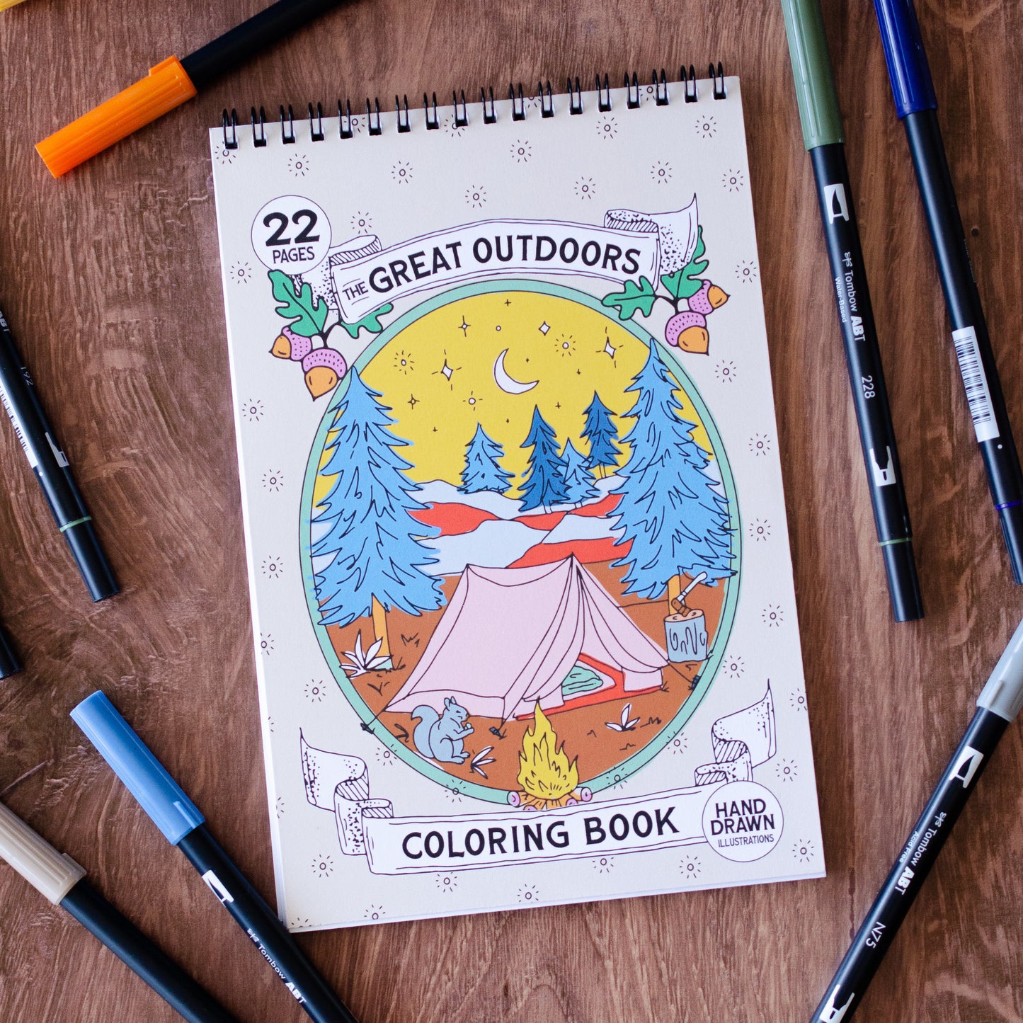 The Great Outdoors Coloring Book with 22 pages of hand-drawn nature illustrations featuring a camping scene, tent, pine trees, wildlife, and a cozy campfire under the stars. Spiral-bound adult coloring book for outdoor and adventure lovers.