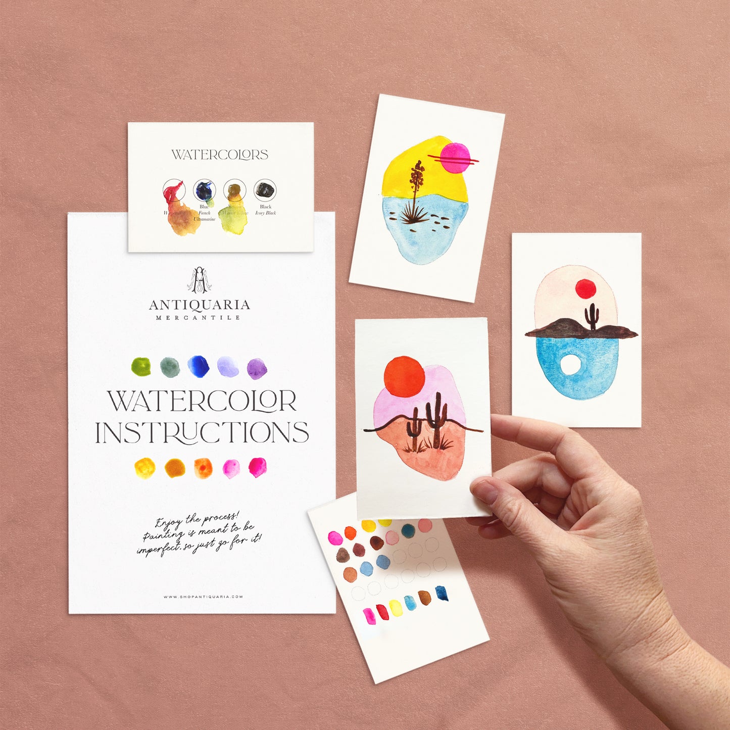 Hand holding a watercolor instruction card with colorful designs on a pink background