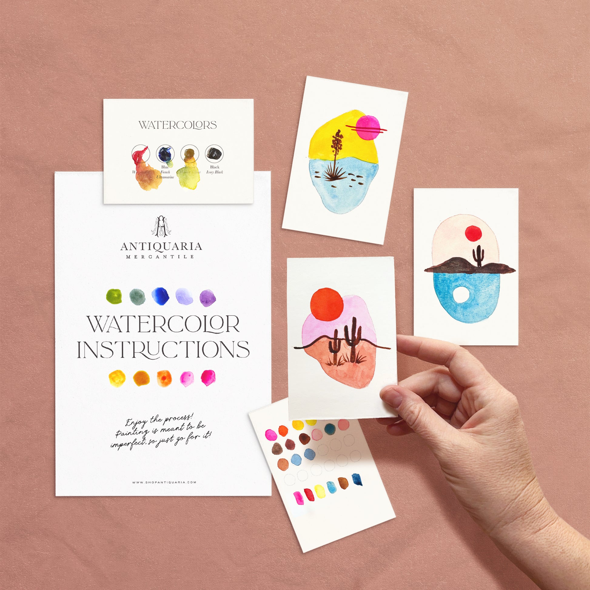 Hand holding a watercolor instruction card with colorful designs on a pink background